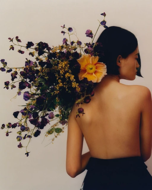 Bare-backed woman with short dark hair, carrying bouquet of purple and yellow flowers against white wall, studio shot.