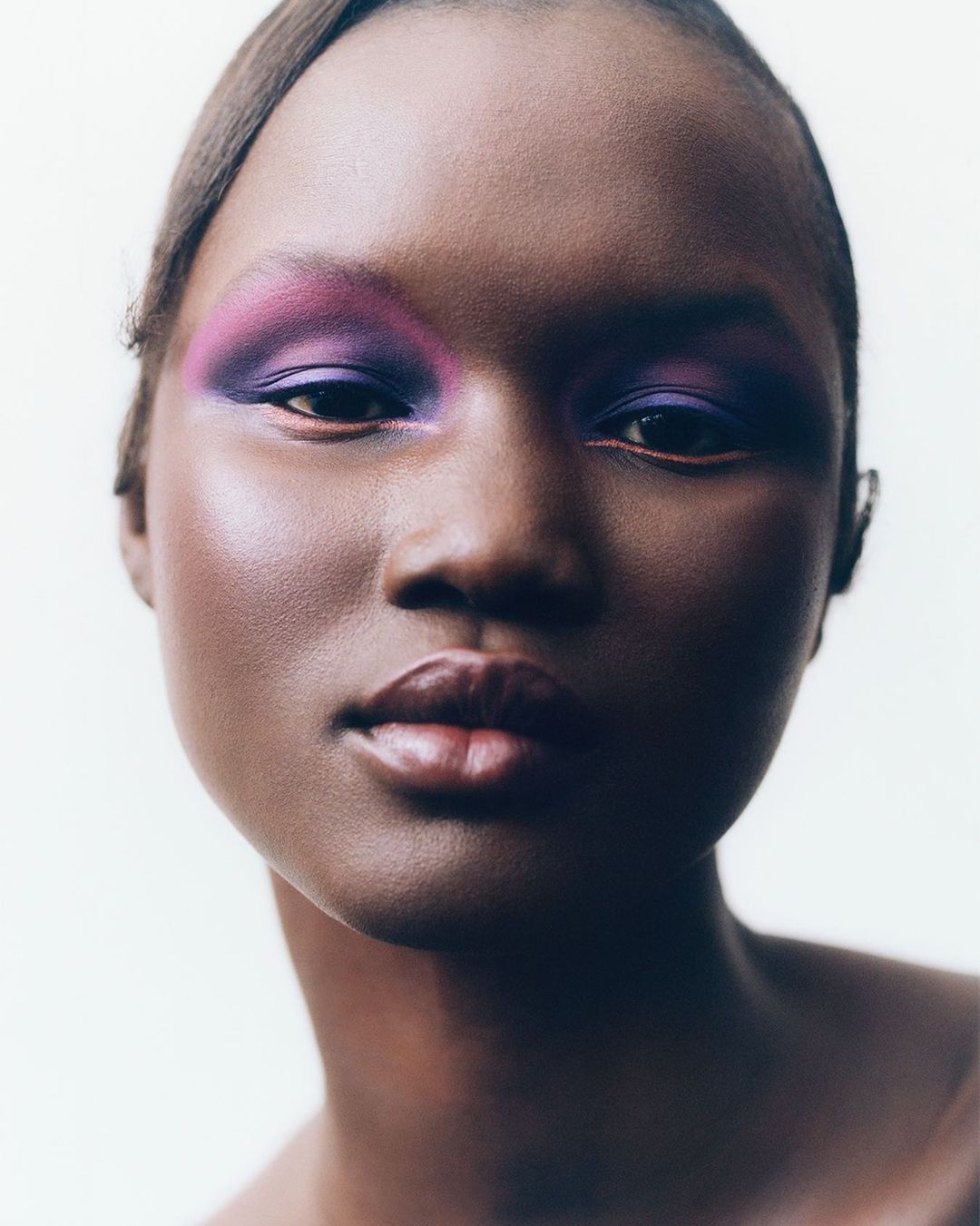 Close-up portrait of a Black female model with purple and pink eyeshadow and glossy lips, showcasing a beauty or fashion aesthetic.