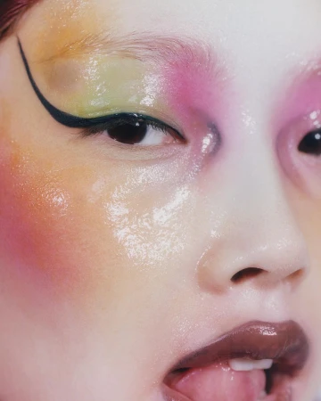 Close-up portrait of a model with glossy pastel yellow and pink eye makeup, winged black eyeliner, and glossy dark lips by Felicity Ingram.