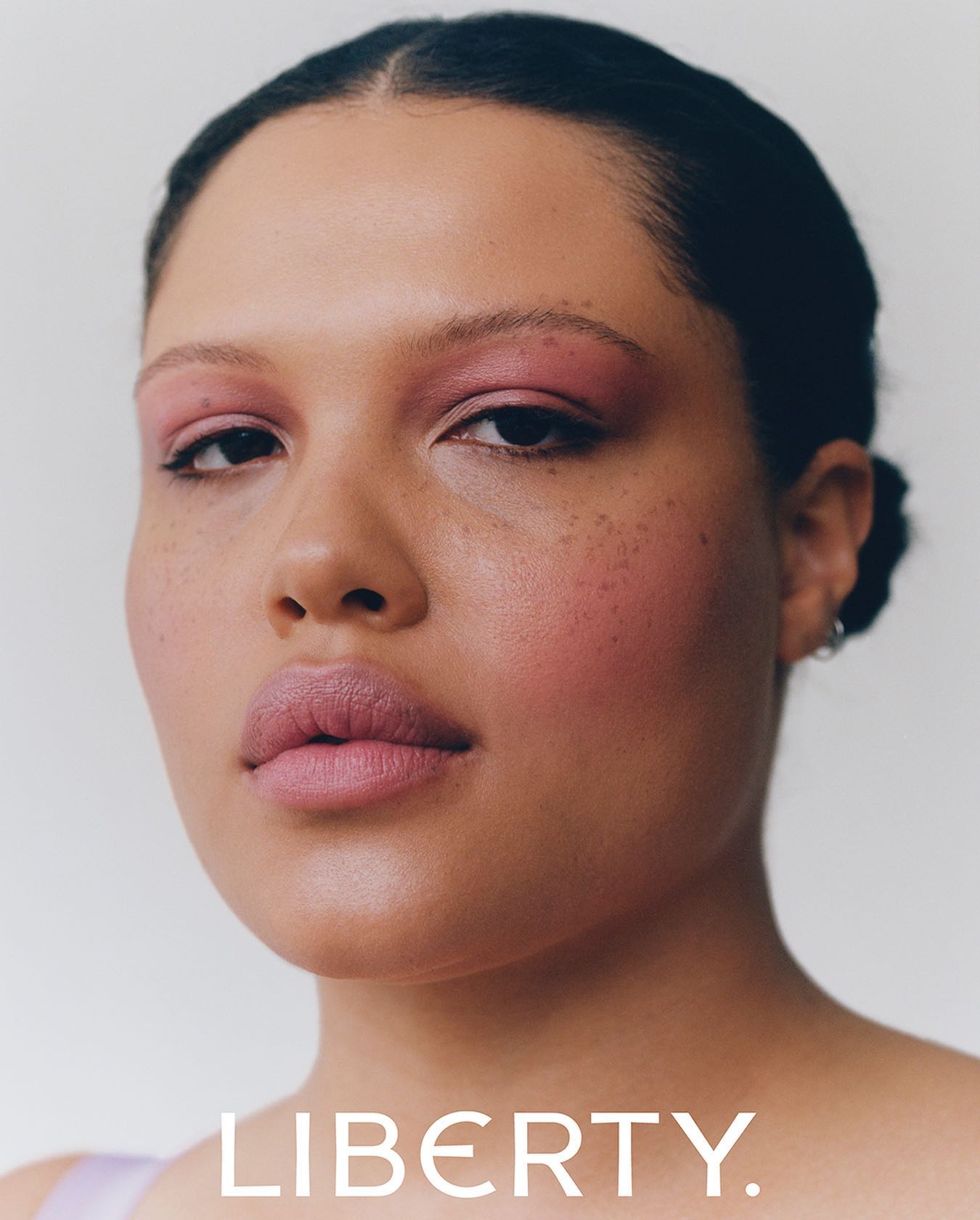 Close-up portrait of an adult female model with pink eye shadow and lipstick against a clean white backdrop, showcasing beauty and makeup.