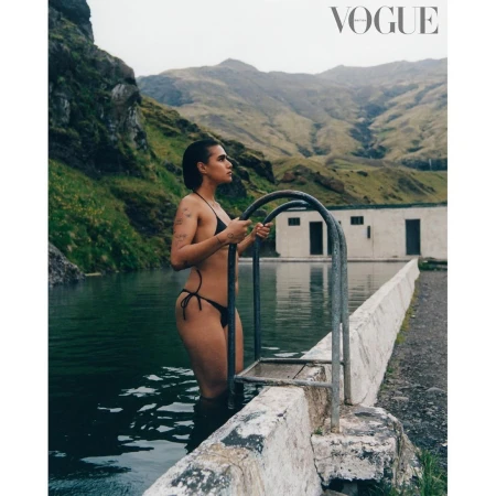 Woman in a black bikini exiting a geothermal pool in a mountainous region, with a building and Vogue logo visible.