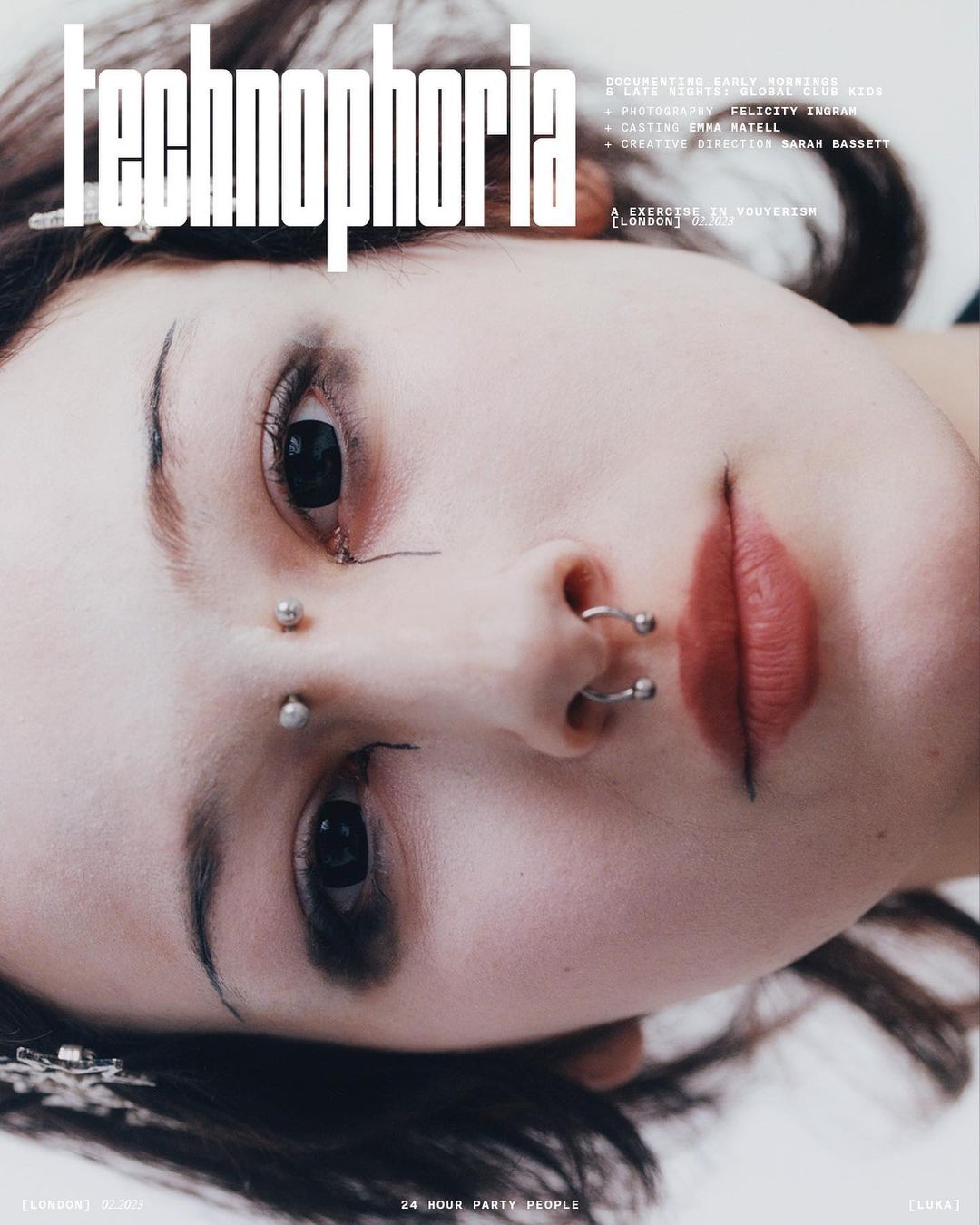 Close-up portrait of a young adult woman with multiple facial piercings. She has fair skin, dark hair, and distinct eyebrow makeup.