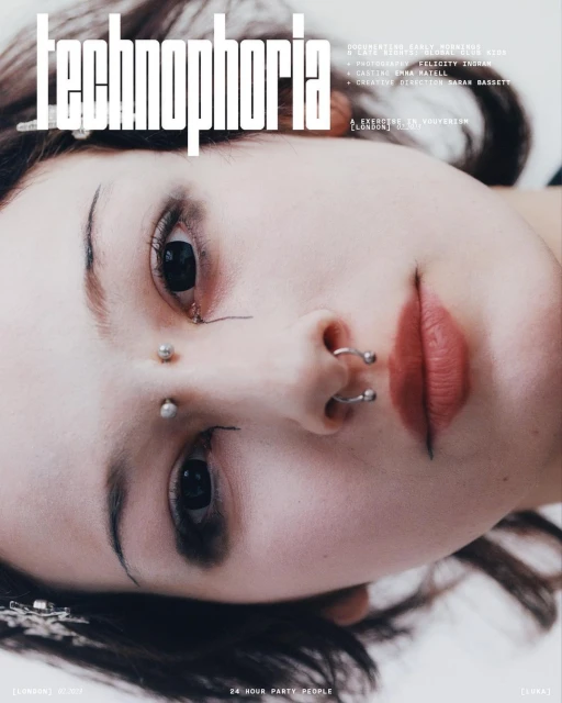 Close-up portrait of a young adult woman with multiple facial piercings. She has fair skin, dark hair, and distinct eyebrow makeup.