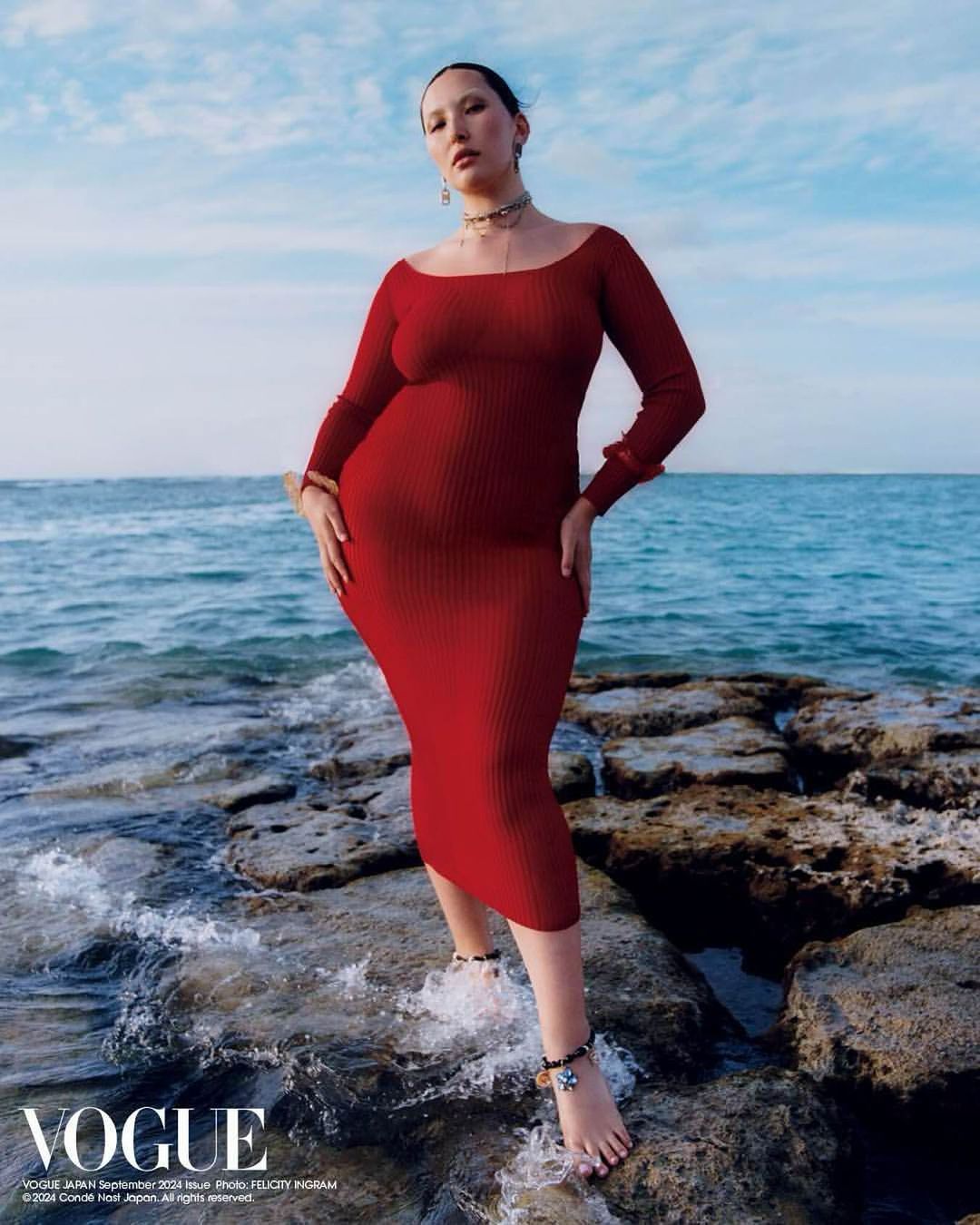 Woman in a red long-sleeve dress poses barefoot on rocks at the shoreline with the ocean and blue sky in the background.