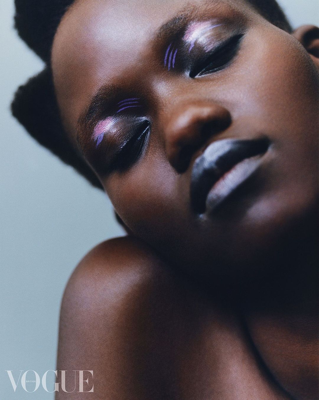 Close-up fashion shot of a dark-skinned woman with creative white and purple makeup, eyes closed, against a light blue background.
