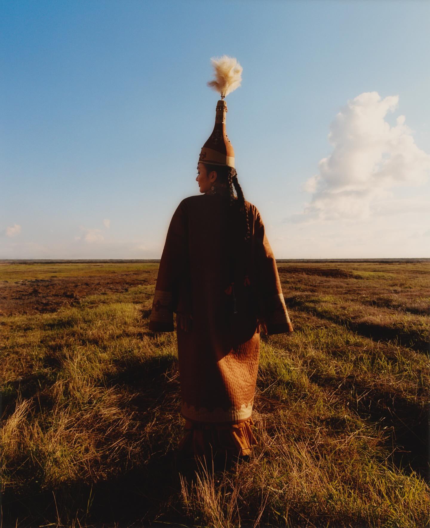 Woman in traditional clothing and headdress stands in a grassy field under a blue sky, facing away from the viewer in a portrait photo.