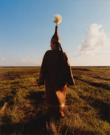 Woman in traditional clothing and headdress stands in a grassy field under a blue sky, facing away from the viewer in a portrait photo.
