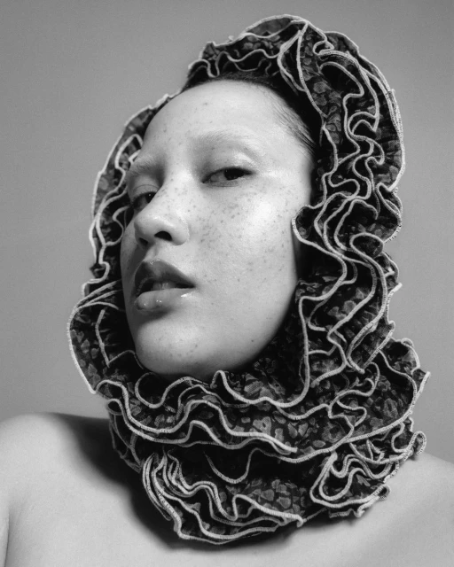 Monochrome portrait of a woman with freckles wearing a high-ruffled, floral lace collar and neutral makeup in a studio setting.