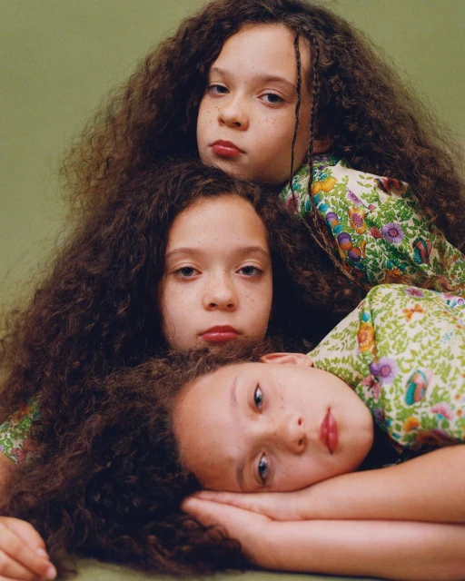 Three young girls with dark curly hair wearing floral tops stacked on each other against a green backdrop in a studio portrait.