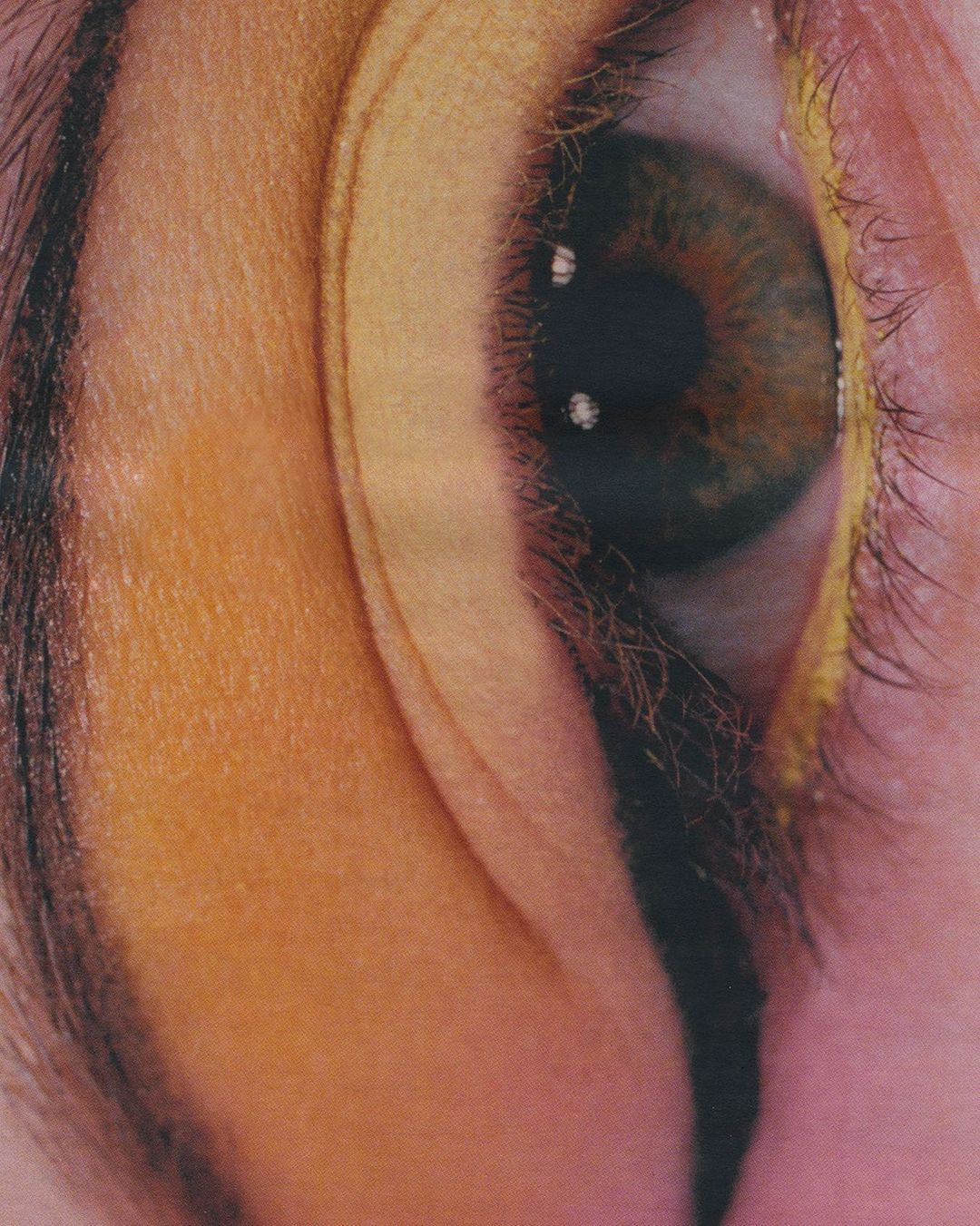Close-up of a human eye with brown iris and yellow eyeshadow. The makeup is thick and exaggerated, drawing attention to the eye.