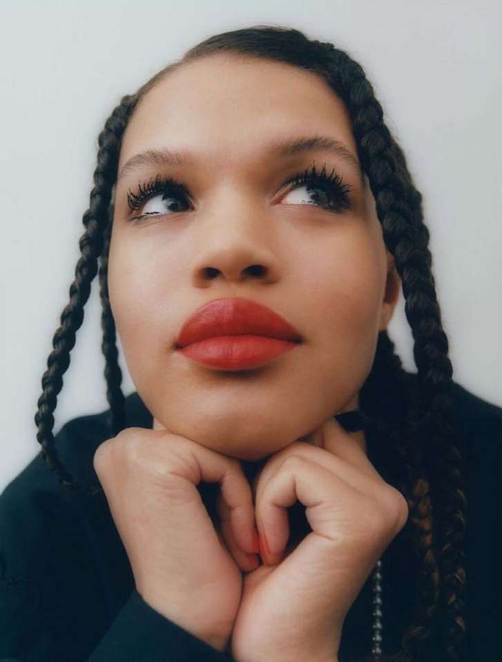 Close-up portrait of a young adult woman with braided dark hair, red lipstick, and her hands gently supporting her chin.