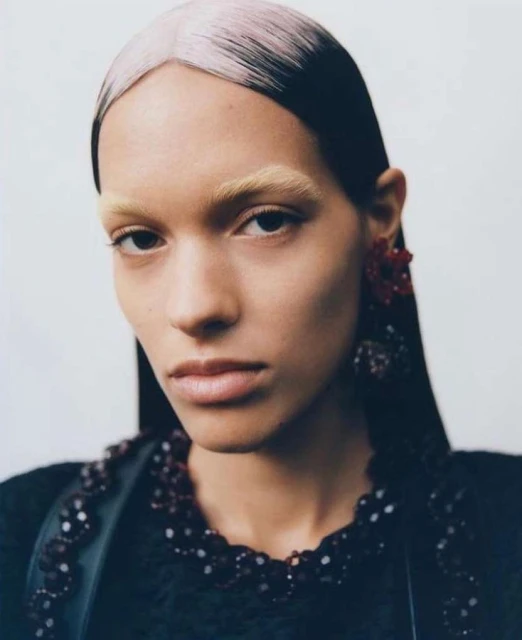 Close-up studio shot of a fashion model with a serious expression wearing a beaded black top and earrings, highlighted by a stark white background.
