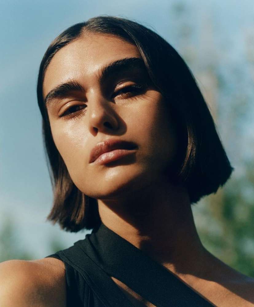Close-up portrait of a woman with short dark hair and dark eyebrows, wearing a black top, posing outdoors against a blue and green backdrop.