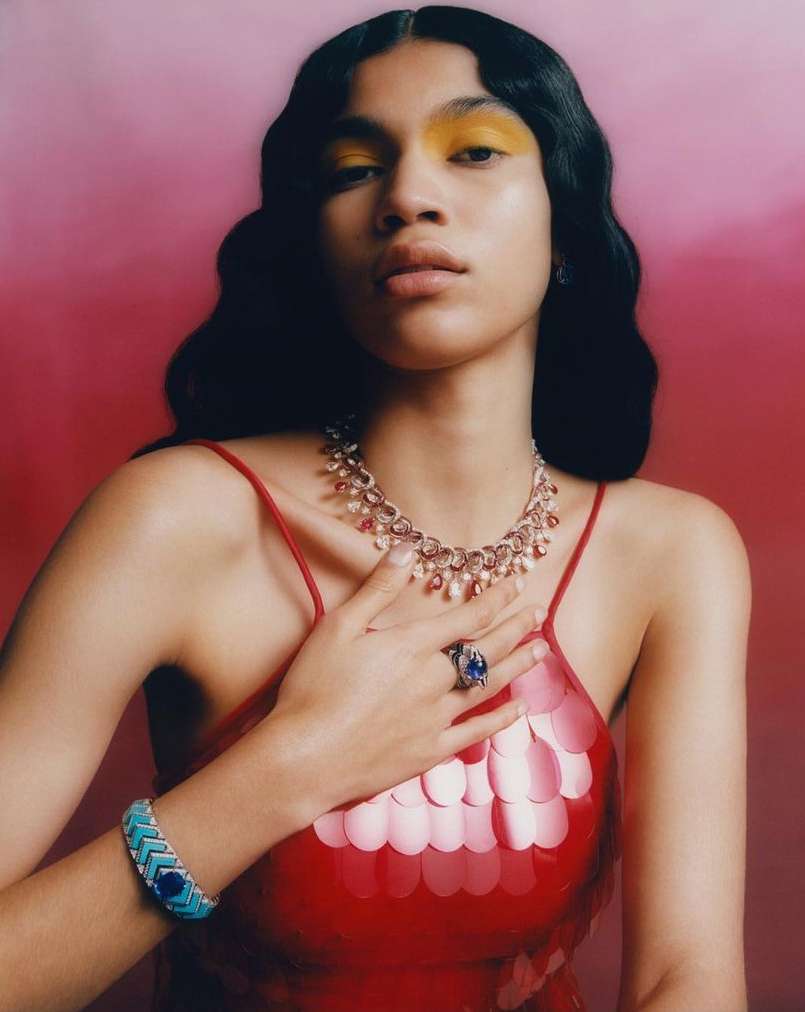 A model wears red clothing with jewelry including a necklace, bracelet, and ring against a red and pink background in a studio shot.