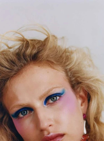 Close-up portrait of a blonde woman with dramatic blue eye makeup and pink blush. Red and silver earrings are visible against a white background.