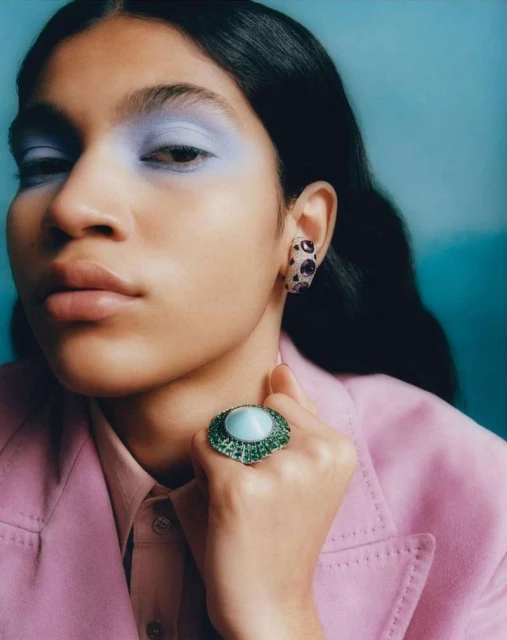 Close-up of a person wearing jewelry and makeup, showcasing a large ring and stylish earrings, against a blurred turquoise backdrop.