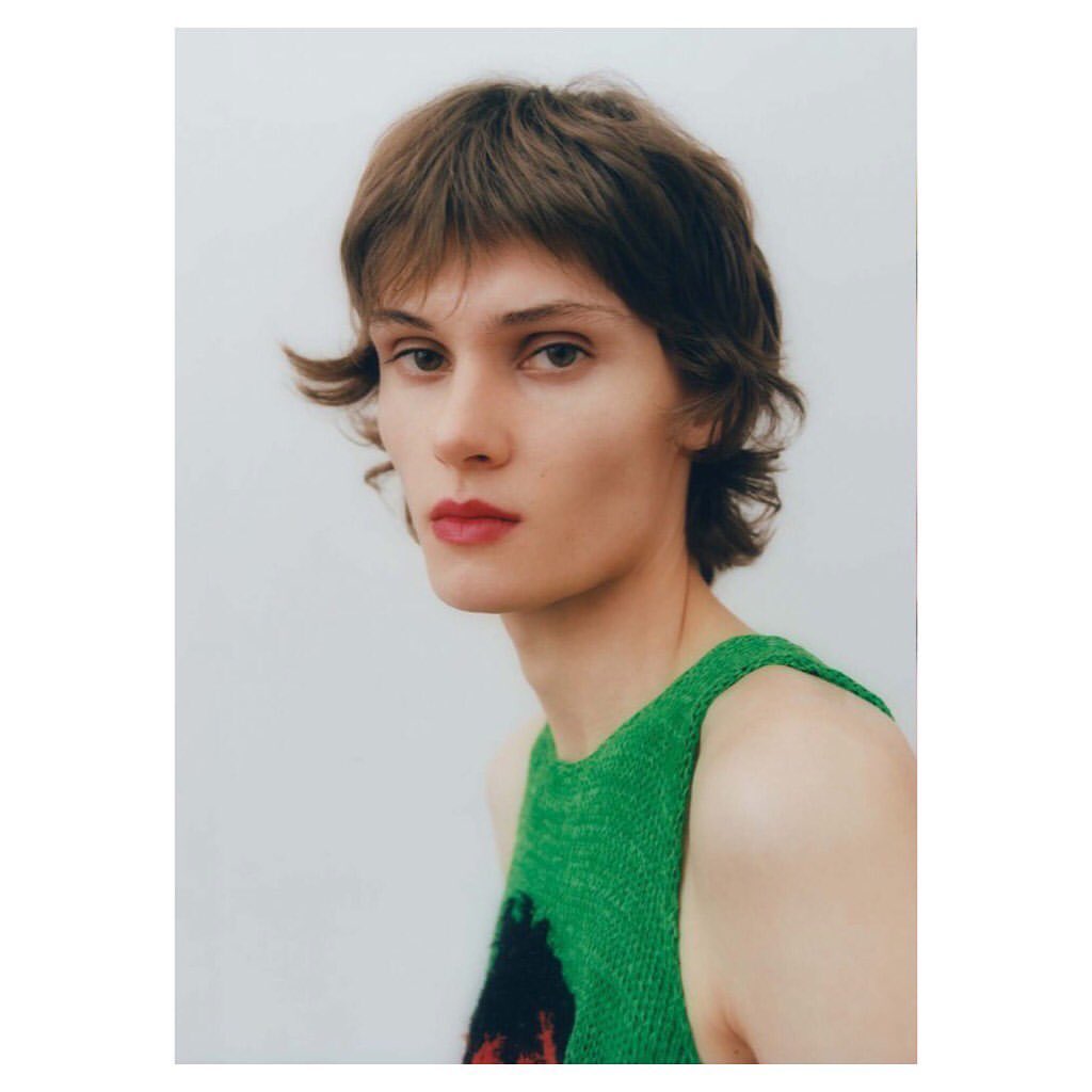Portrait of androgynous adult female with short brown hair, wearing a green and black patterned tank top, against a white background.