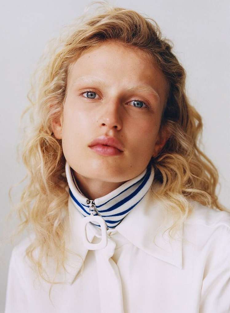 Close-up portrait of a blonde woman with blue eyes, wearing a white shirt and a striped collar, set against a bright background.