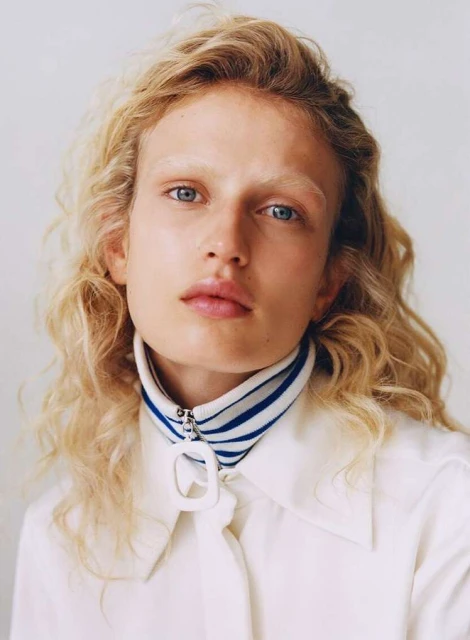 Close-up portrait of a blonde woman with blue eyes, wearing a white shirt and a striped collar, set against a bright background.