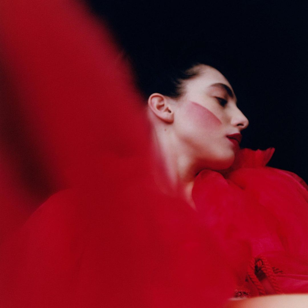 Profile portrait of a fair-skinned woman with red makeup and a red tulle collar against a black background in a beauty editorial shot.