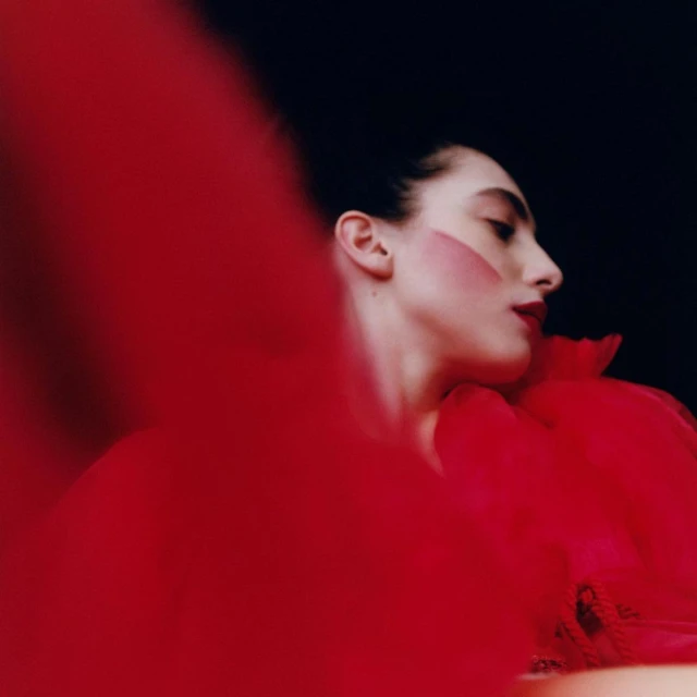 Profile portrait of a fair-skinned woman with red makeup and a red tulle collar against a black background in a beauty editorial shot.