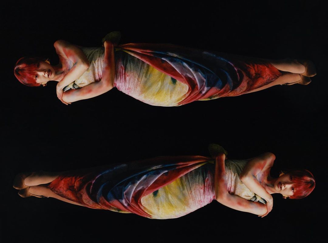 Two young adult women symmetrically posed against a black background, wearing light dresses with rainbow-colored accents, and short red hair.