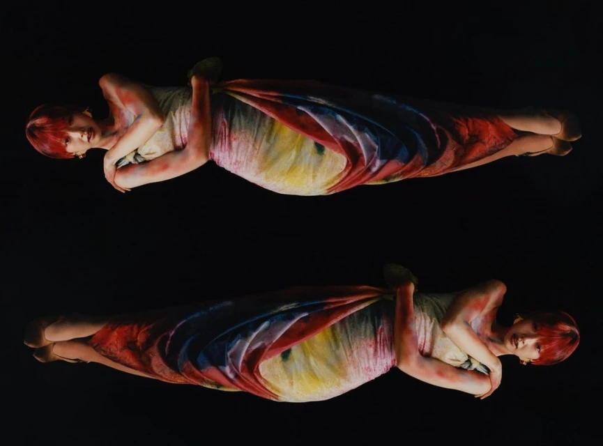 Two young adult women symmetrically posed against a black background, wearing light dresses with rainbow-colored accents, and short red hair.