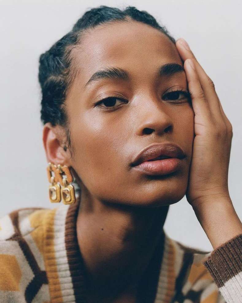 Portrait of an adult woman with dark hair and gold earrings, wearing a patterned sweater against a light background.