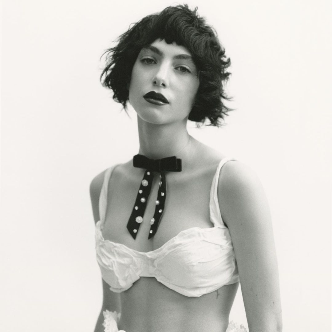 Black and white portrait of a woman with dark hair and red lipstick wearing a white bra top and a velvet bow tie with pearls.