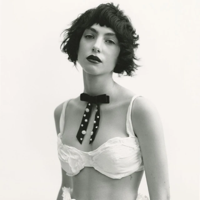 Black and white portrait of a woman with dark hair and red lipstick wearing a white bra top and a velvet bow tie with pearls.