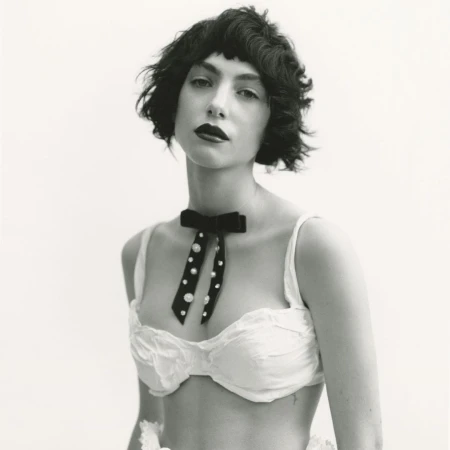 Black and white portrait of a woman with dark hair and red lipstick wearing a white bra top and a velvet bow tie with pearls.