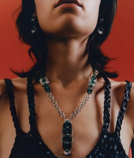 Woman wearing a black braided top and statement emerald jewelry including earrings and a necklace in a studio setting with a red background.