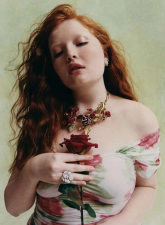 Red-haired woman poses with a red rose, wearing a floral dress and rose-themed jewelry against a muted green backdrop in a close-up shot.