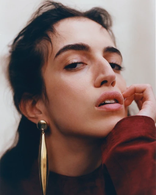 Close-up portrait of a young woman with dark hair and gold drop earrings, wearing a red top against a neutral background.