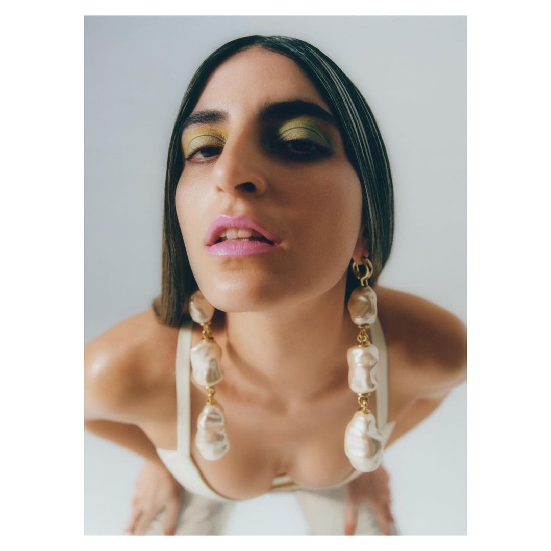 Woman with long dark hair wearing statement pearl earrings and bright makeup, low-angle close-up in photo studio against a plain white background.