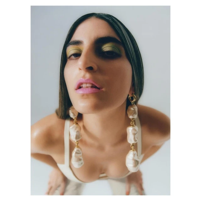 Woman with long dark hair wearing statement pearl earrings and bright makeup, low-angle close-up in photo studio against a plain white background.