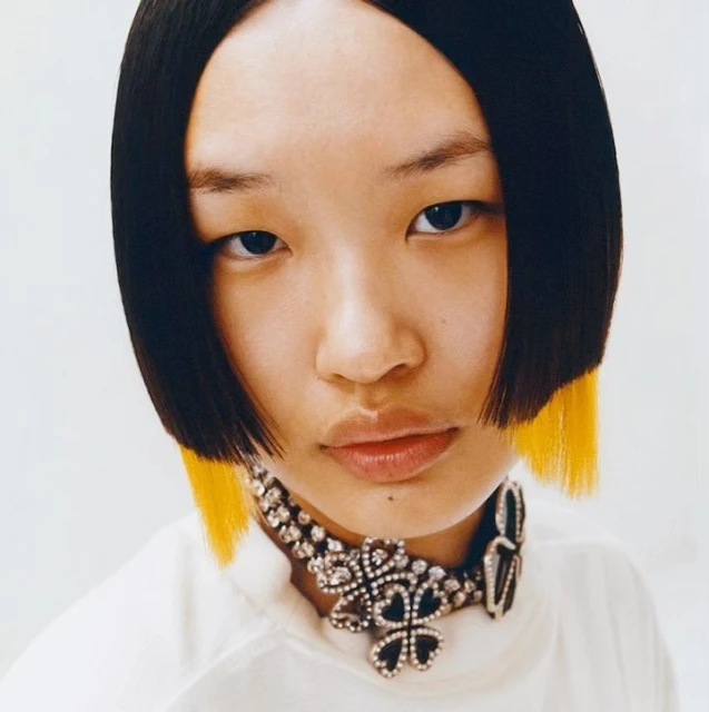 Close-up portrait of an Asian woman with a blunt black bob haircut with yellow tips, wearing a decorative jeweled choker necklace.