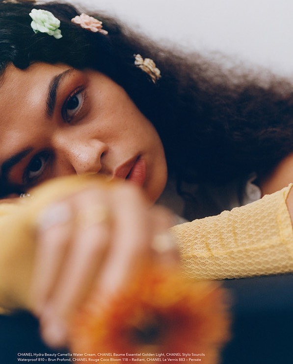 Portrait of an adult female with dark curly hair, adorned with small fabric flowers, resting her head on arms, with a flower in foreground.