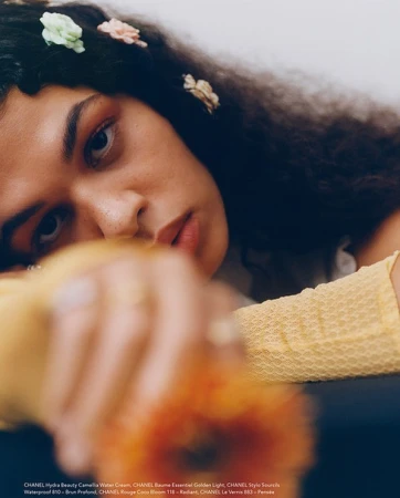 Portrait of an adult female with dark curly hair, adorned with small fabric flowers, resting her head on arms, with a flower in foreground.