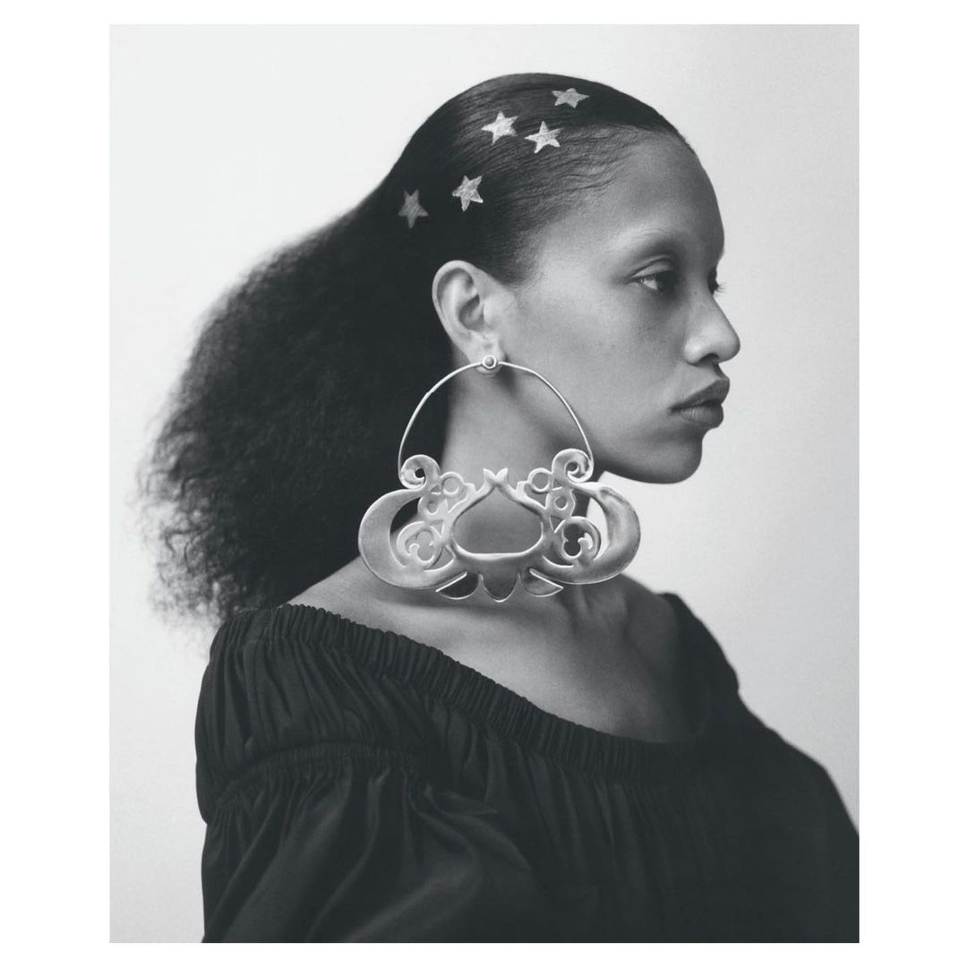 Portrait of a woman with dark hair adorned with star-shaped accents, showcasing large, ornate silver hoop earrings against a bright backdrop.
