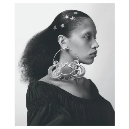 Portrait of a woman with dark hair adorned with star-shaped accents, showcasing large, ornate silver hoop earrings against a bright backdrop.