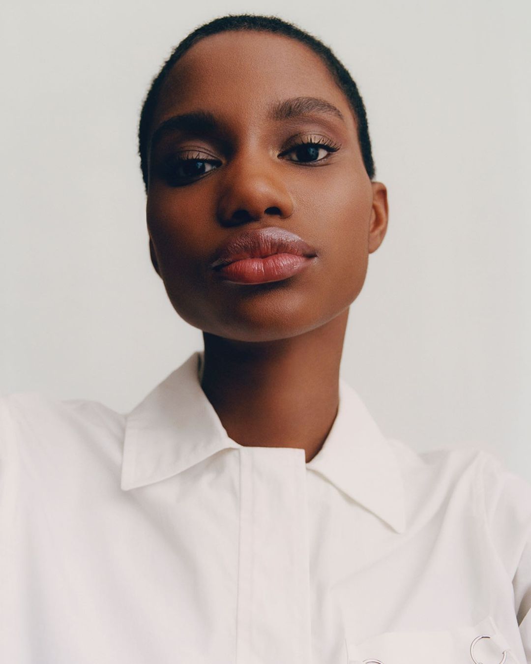 Close-up portrait of a Black woman with short hair wearing a white collared shirt against a white background, gazing directly at the camera.