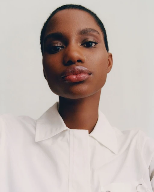 Close-up portrait of a Black woman with short hair wearing a white collared shirt against a white background, gazing directly at the camera.