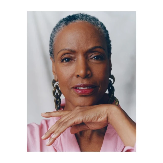 Portrait of a smiling, grey-haired Black woman with large earrings, wearing a pink shirt and posing in front of a white backdrop.