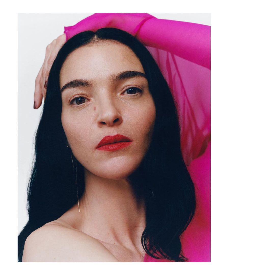 Close-up portrait of a woman with dark hair and bright red lipstick wearing a sheer hot pink top against a plain white background.
