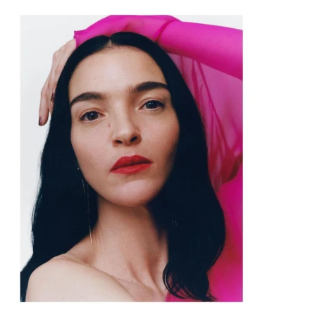 Close-up portrait of a woman with dark hair and bright red lipstick wearing a sheer hot pink top against a plain white background.