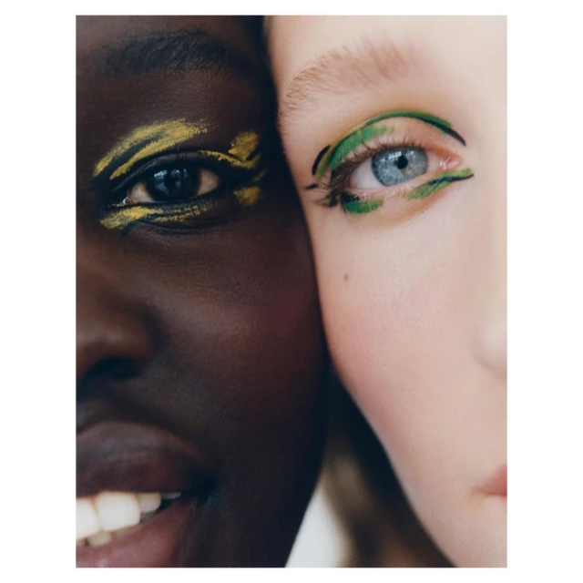 Close-up of two adult females with dramatic green and yellow makeup highlighting their eyes, fair and dark skin tones together.
