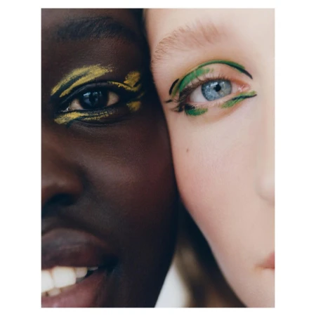 Close-up of two adult females with dramatic green and yellow makeup highlighting their eyes, fair and dark skin tones together.