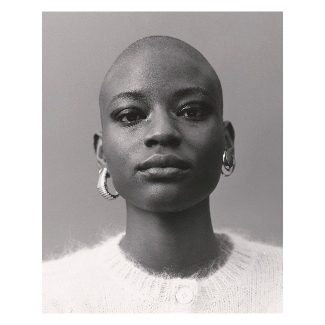 Portrait of a young Black woman with a shaved head, wearing a white sweater and hoop earrings, posing in front of a neutral background.