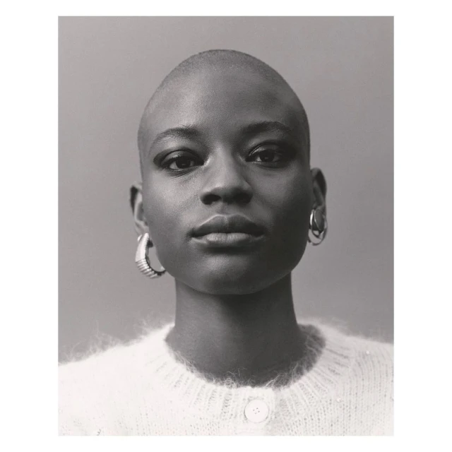 Portrait of a young Black woman with a shaved head, wearing a white sweater and hoop earrings, posing in front of a neutral background.